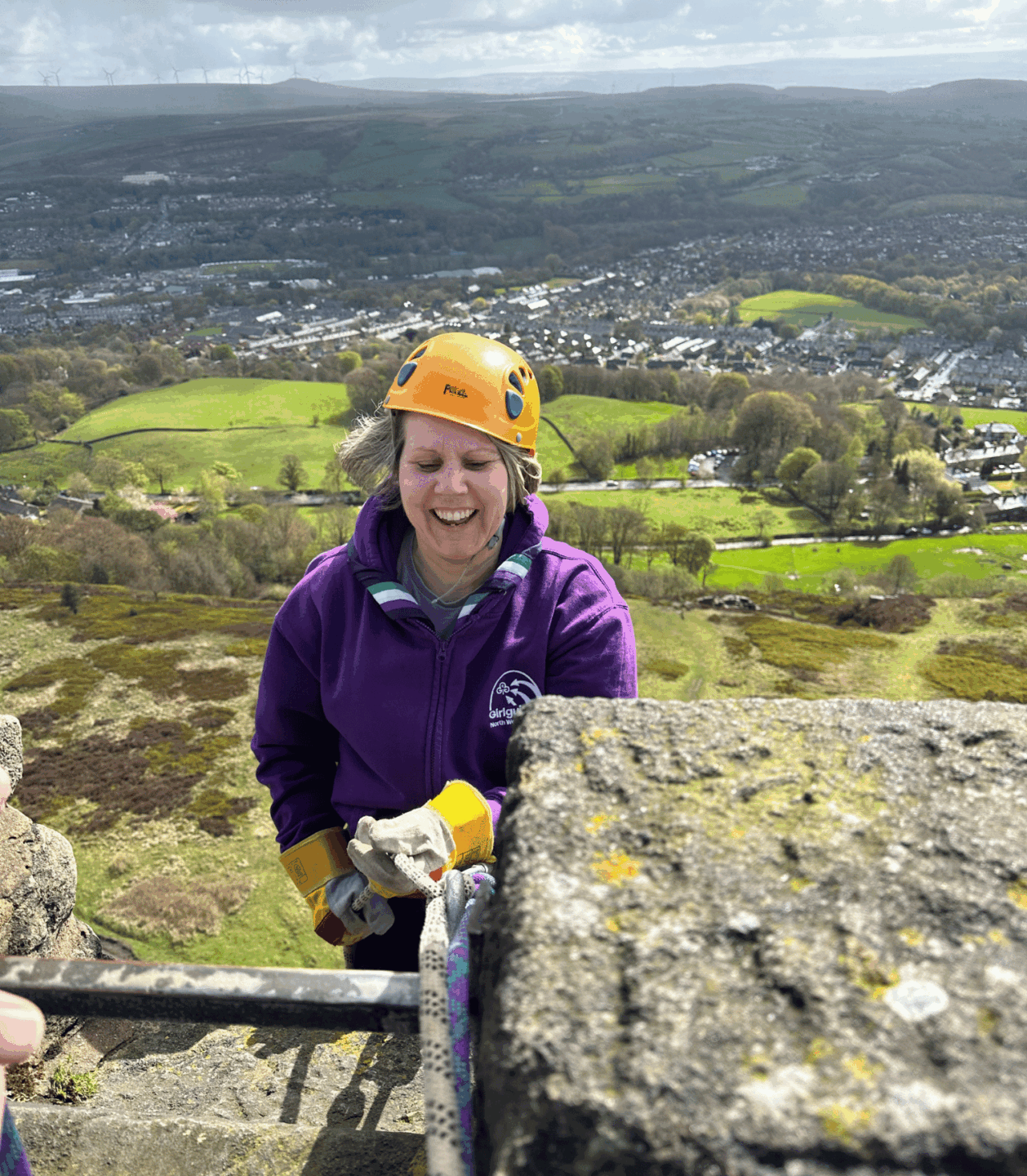 A member of the GGNWE abseils from the top of Peel Tower as part of a fundraiser. They are wearing an orange climbing helmet and a purple region branded hoody.